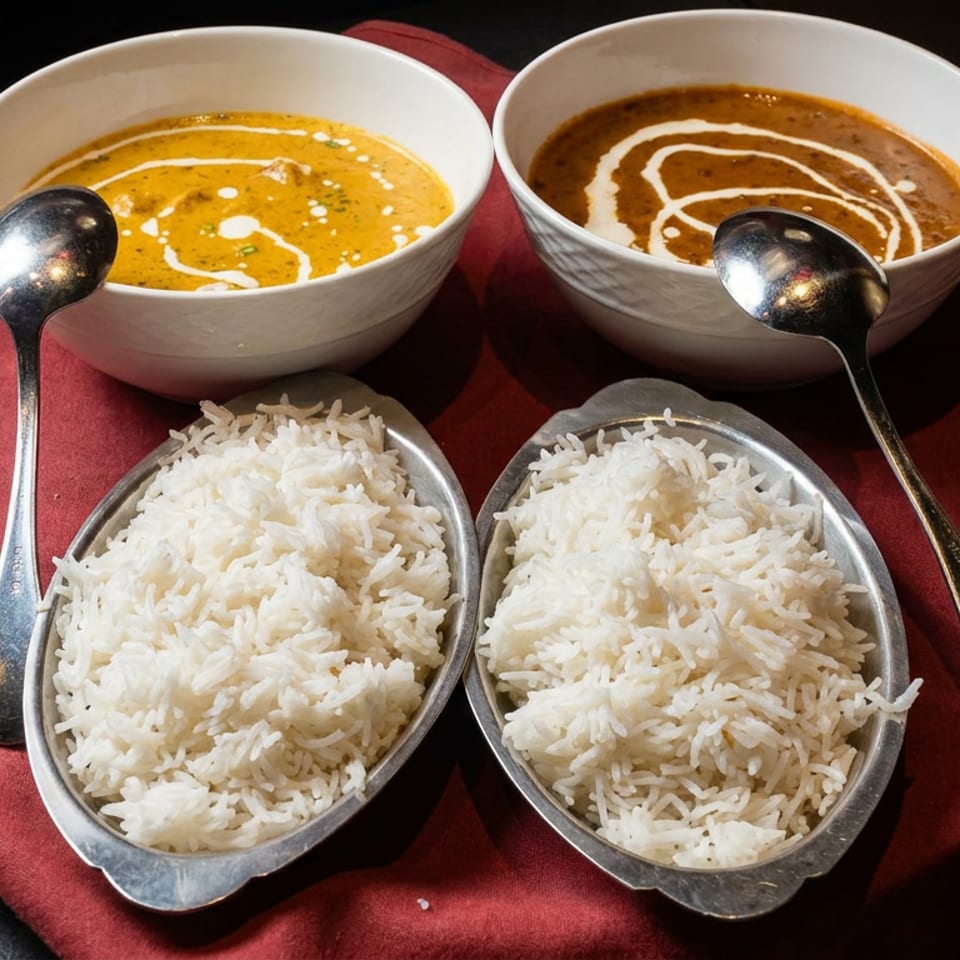 Two bowls of creamy curry topped with white sauce swirls sit behind two serving dishes of basmati rice on a red cloth.