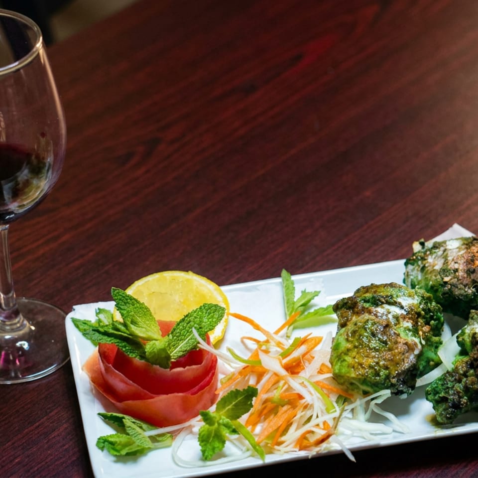 Platter of grilled green herb-coated appetizers with shredded vegetables, lemon, mint, and tomato beside a glass of red wine.